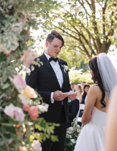 A groom in a tuxedo reads vows to the bride in a white dress during an outdoor wedding ceremony under trees.