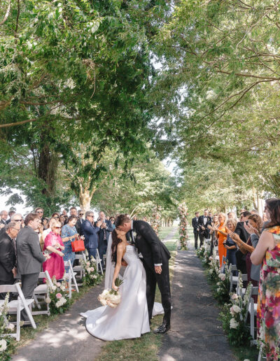 A bride and groom kiss at an outdoor wedding ceremony with guests seated on either side, surrounded by green trees.