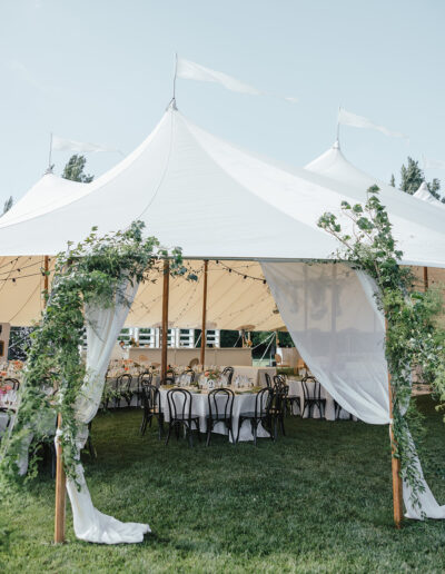 A large white tent with greenery and white fabric draping at the entrance stands on a grassy area, housing tables and black chairs set for an event.