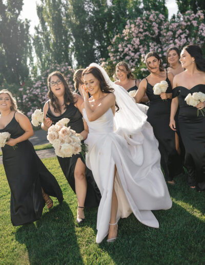 Bride in a white gown walks with bridesmaids in black dresses holding white bouquets in a garden setting.