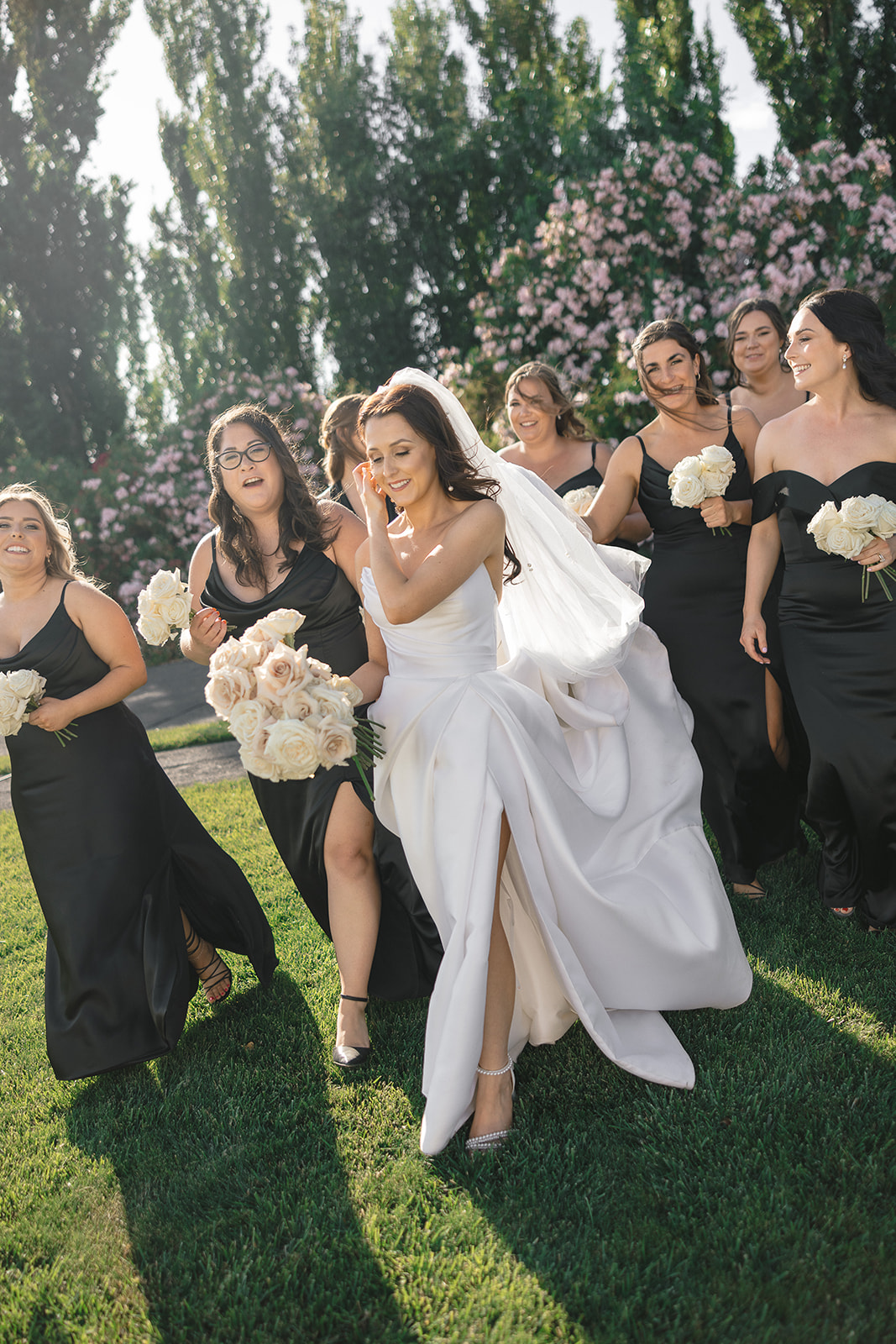 Bride in a white gown walks with bridesmaids in black dresses holding white bouquets in a garden setting.