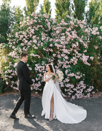 A bride in a white dress and a groom in a black suit stand in front of a large bush with pink flowers.