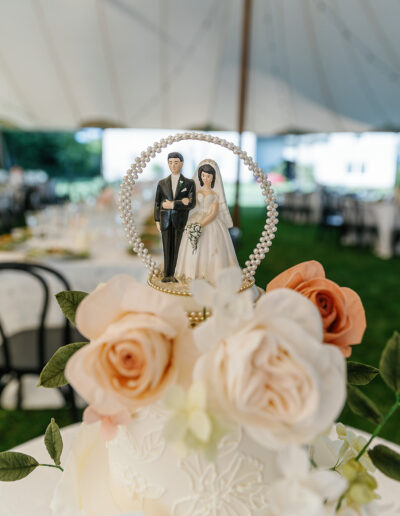 A wedding cake topped with a bride and groom figurine is adorned with large cream and peach roses, under a white tent at an outdoor event.