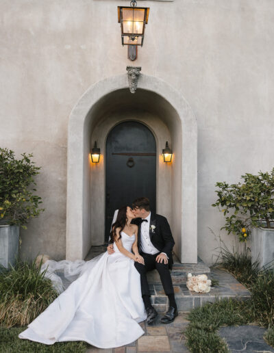 A bride and groom sit and kiss on steps in front of an arched door, with soft outdoor lighting and two potted plants nearby.