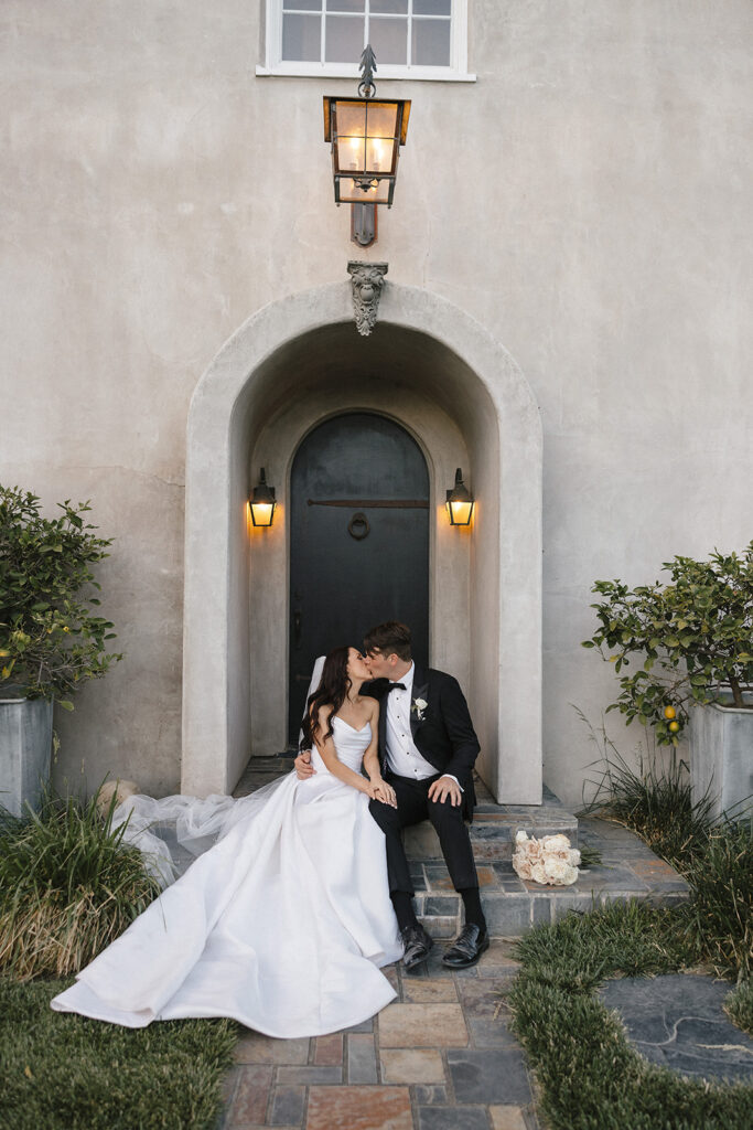 A bride and groom sit and kiss on steps in front of an arched door, with soft outdoor lighting and two potted plants nearby.