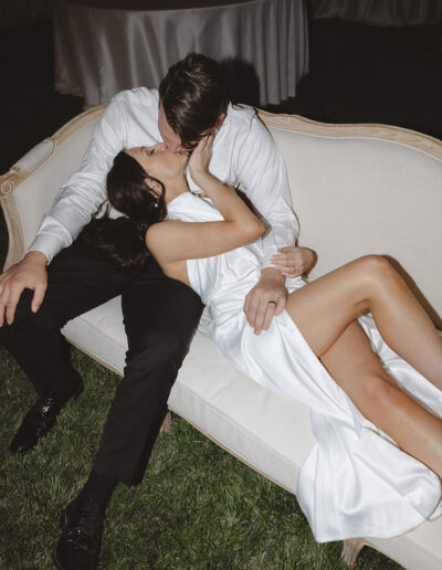 A couple in formal attire is sitting on a sofa outdoors, kissing. The woman is wearing a white dress and the man is in a white shirt and black pants.