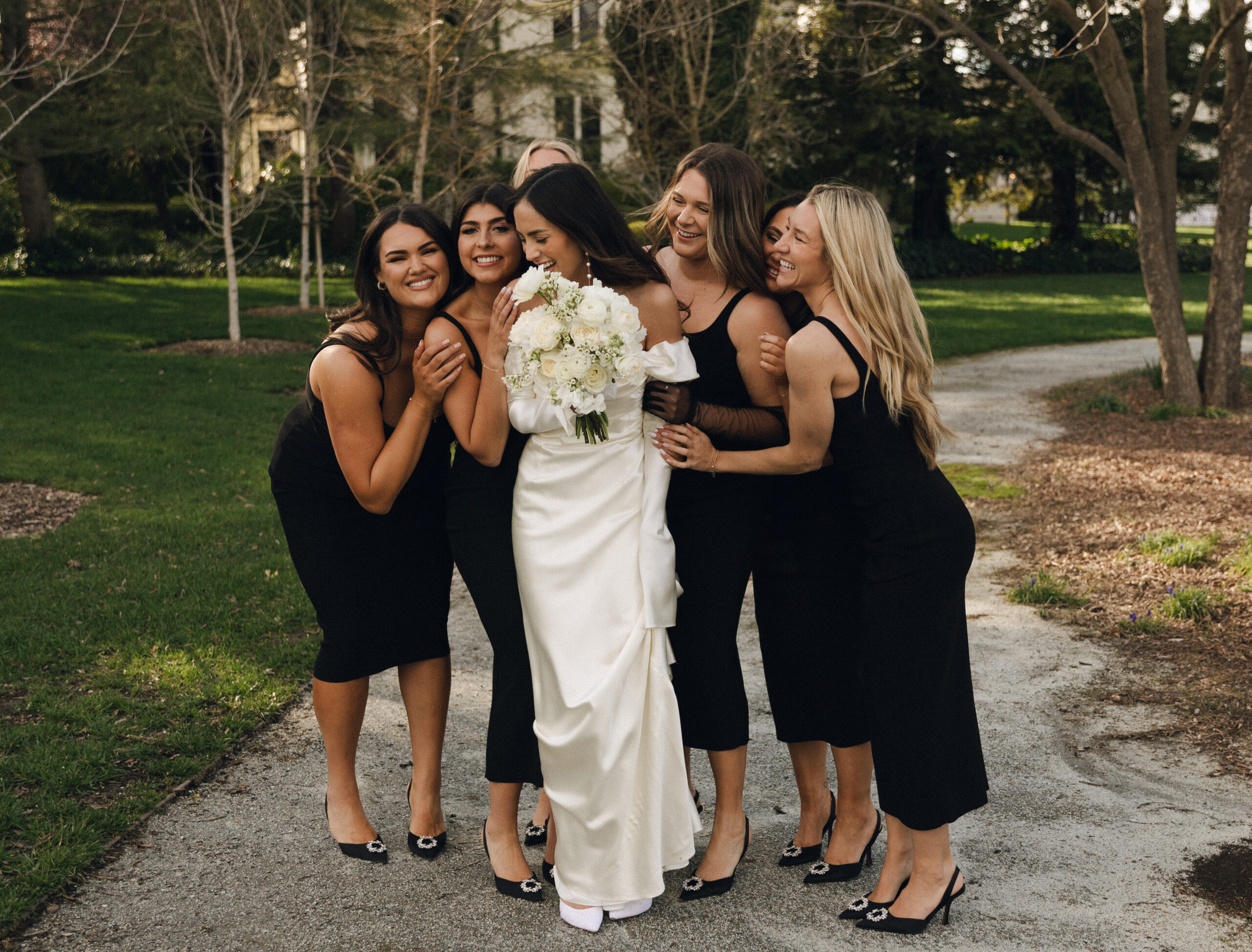 A bride in a white dress holds a bouquet and is surrounded by five bridesmaids in black dresses, all standing on a garden path.