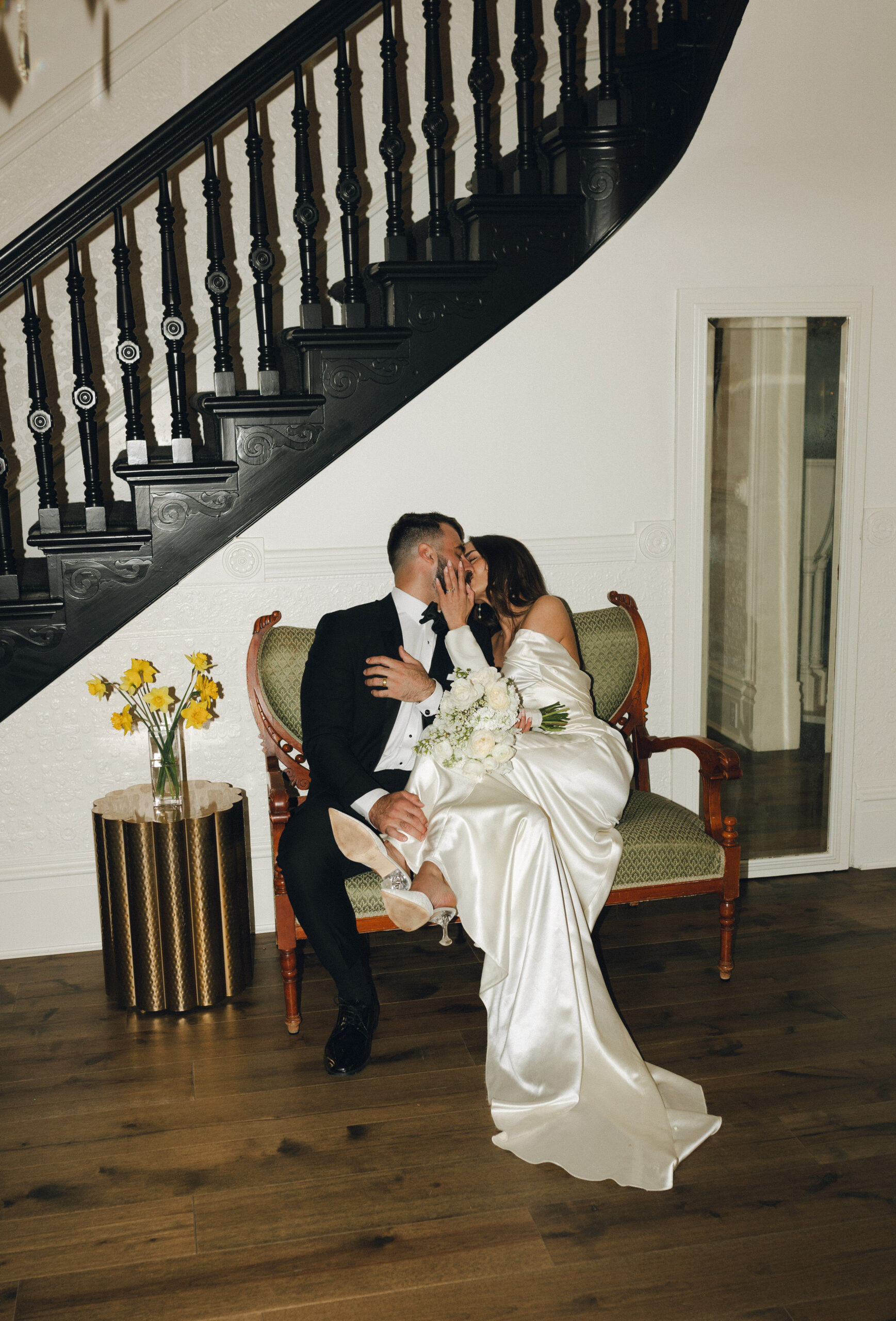 A couple in formal attire kisses while seated on a green upholstered bench. A spiral staircase is in the background, and a small table with daffodils is on the left.