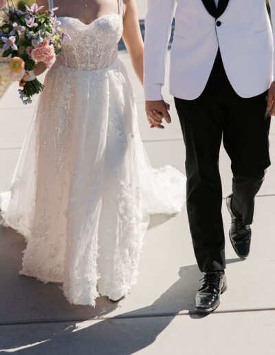 A bride in a white dress and a groom in a white jacket and black pants hold hands while walking on a path, with the bride carrying a bouquet of flowers.