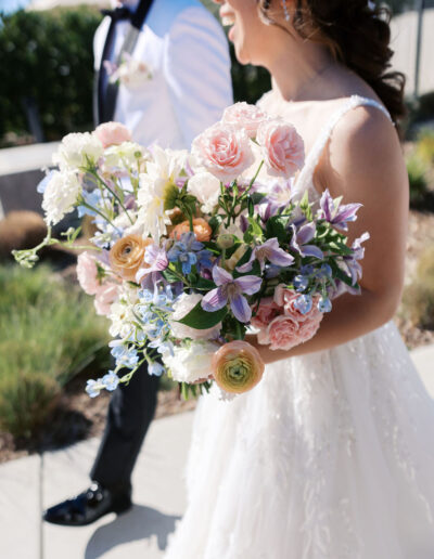 A bride in a white gown holds a large bouquet of pink, white, and purple flowers and walks beside a man in a white jacket and black pants.