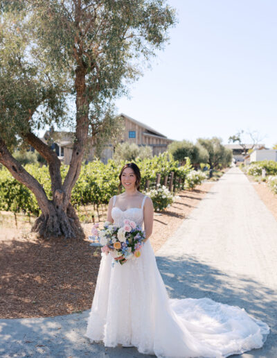 Bride in a white gown stands on a dirt path, holding a flower bouquet. Vineyards and trees are in the background.