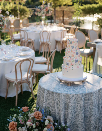 A multi-tiered floral cake on a decorated table in an outdoor setting, surrounded by elegantly set tables and chairs with floral arrangements.