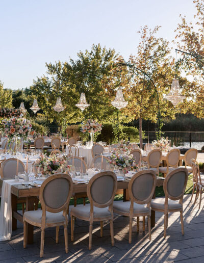 Elegant outdoor wedding setup with long tables, floral centerpieces, wooden chairs, and chandeliers, under a canopy of trees with sunlight filtering through.