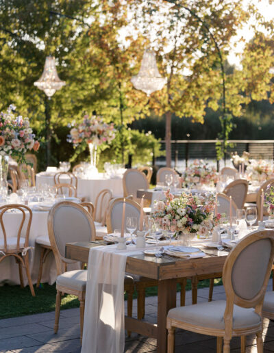 An elegant outdoor event setup with decorated tables, floral centerpieces, and chandelier lights under a canopy of trees.