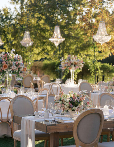 An outdoor wedding reception setup with elegantly decorated tables, floral centerpieces, and overhead chandeliers, set against a backdrop of trees and greenery.