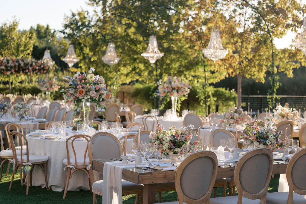 An outdoor wedding reception setup with elegantly decorated tables, floral centerpieces, and overhead chandeliers, set against a backdrop of trees and greenery.