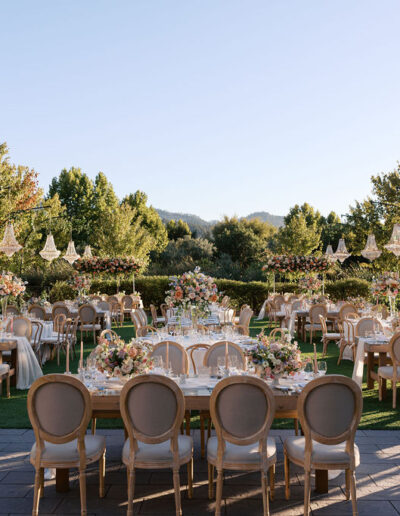 Elegant outdoor wedding setup with round tables, floral arrangements, chandeliers, and wooden chairs on a grassy area, surrounded by trees under a clear sky.