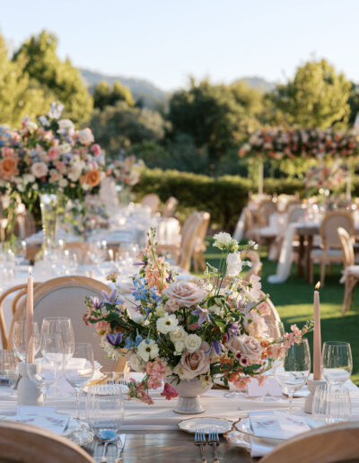 An elegant outdoor dining setup with floral centerpieces, glassware, and neutral-colored candles on tables surrounded by chairs. Lush greenery and trees are visible in the background.