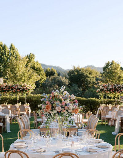 Outdoor wedding reception with round tables, elegant floral centerpieces, and chandeliers, set on green lawn surrounded by trees.