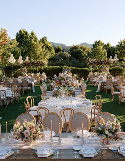 An elegant outdoor wedding reception with tables set for dining, adorned with floral centerpieces and chandeliers. Surrounded by trees on a sunny day.