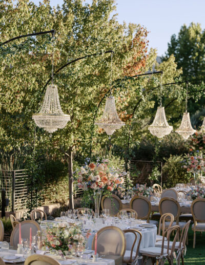 Outdoor wedding reception with tables set for dining, featuring floral arrangements and chandeliers hanging above. Trees and a clear sky form the backdrop.