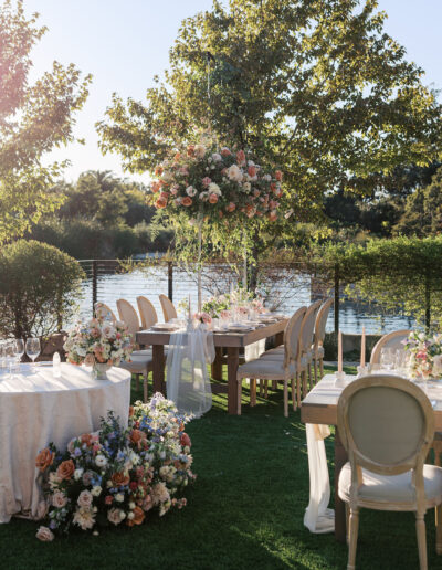 Outdoor wedding setup with decorated tables and chairs on a lawn by a lake. Floral arrangements adorn the tables, and sunlight filters through the trees.