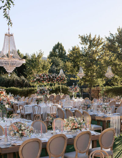 Outdoor wedding reception with elegantly set tables, floral centerpieces, and chandeliers hanging from above. Trees and greenery surround the area.