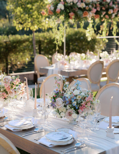 Elegant outdoor dining table setup with floral centerpieces, candles, and place settings. Surrounded by greenery and softly lit by natural sunlight.