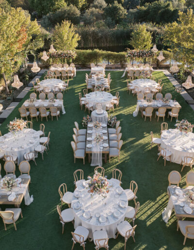 Outdoor wedding reception setup with round and rectangular tables covered in white tablecloths, surrounded by chairs. Tables are decorated with floral centerpieces, set on a grassy area.