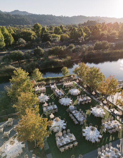 Aerial view of an outdoor event setup with round tables and chairs on a grassy area beside a lake, surrounded by trees and greenery.