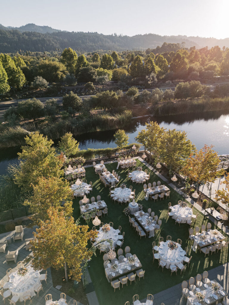 Aerial view of an outdoor event setup with round tables and chairs on a grassy area beside a lake, surrounded by trees and greenery.