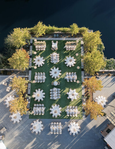 Aerial view of an outdoor event setup with round tables and chairs on a green lawn, surrounded by trees and near a body of water.