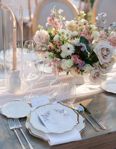 Elegant table setting with floral centerpiece, white plates, crystal glassware, and gold cutlery on a wooden table. Tall candle in a glass holder beside the arrangement.