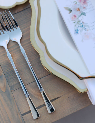 Place setting with two forks, scalloped white plates with gold edges, and a floral menu on a wooden table.