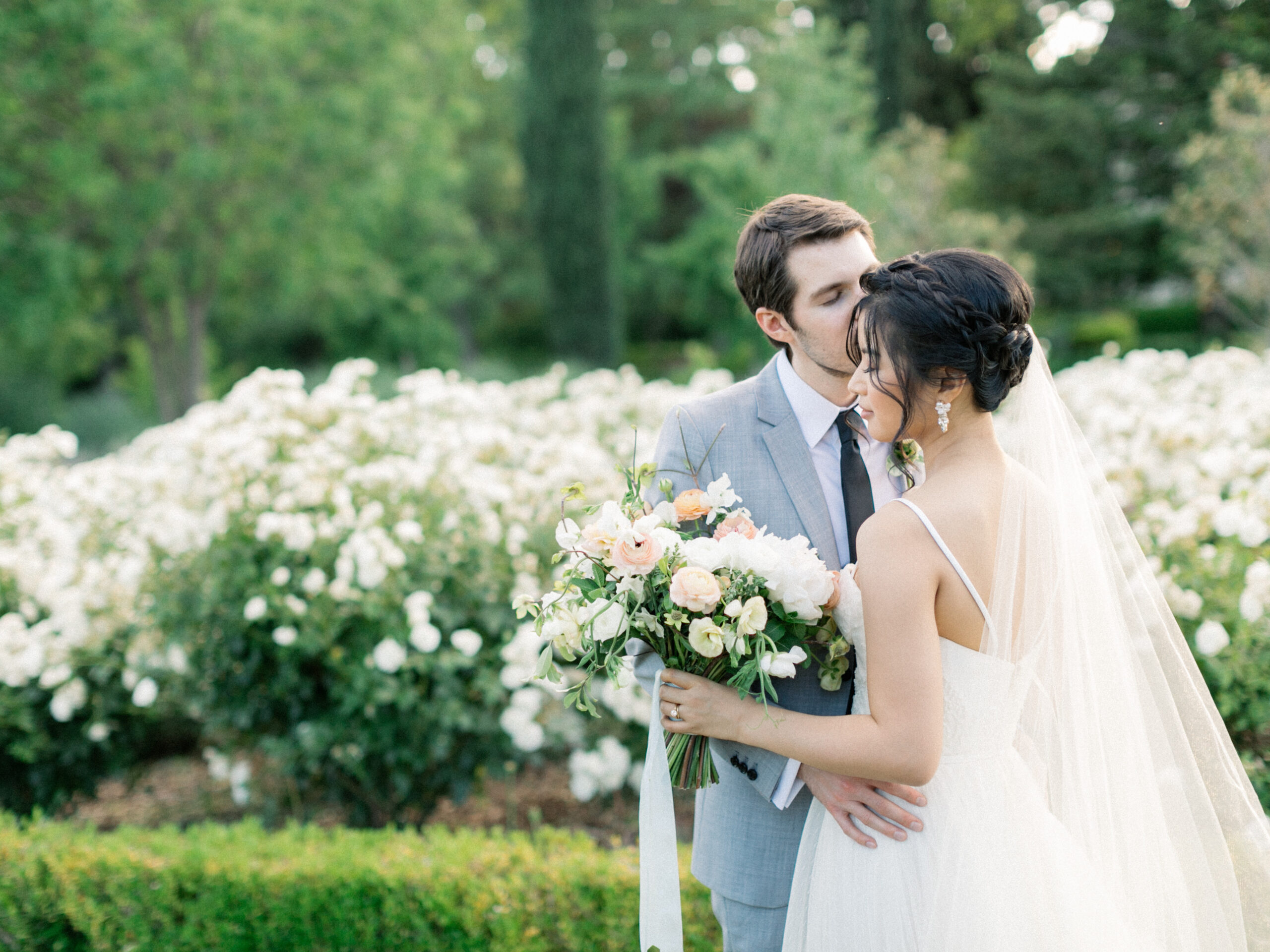 A groom in a light gray suit kisses the bride on the forehead as she holds a bouquet of flowers; they stand in front of blooming white bushes outdoors, perfectly styled by Park Winters Wedding Planner.