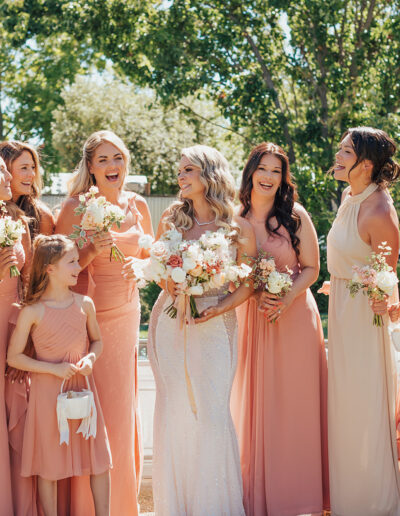 A bride stands with her bridesmaids, all dressed in peach and beige dresses, holding floral bouquets, outdoors on a sunny day.