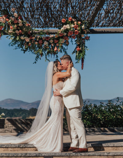 A couple in wedding attire kisses under a floral arch with a mountainous backdrop.