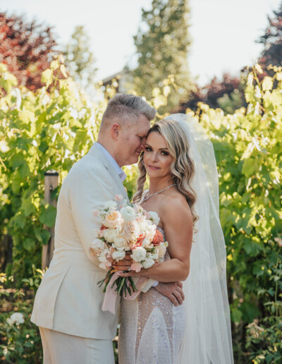 Bride and groom embrace in a vineyard, with the groom in a white suit and the bride holding a bouquet of flowers.