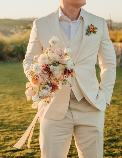 A person in a light-colored suit holds a floral bouquet, standing on grass with a blurred landscape in the background.