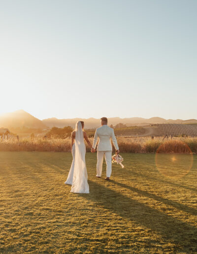 A couple in wedding attire walks hand in hand across a grassy field at sunset.