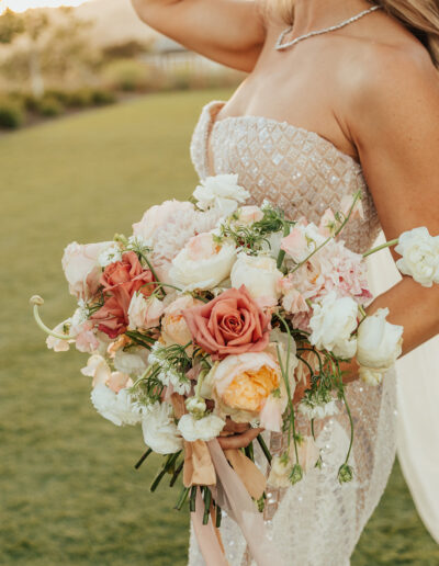 A bride holds a bouquet of pink, peach, and white flowers. She wears a strapless beaded gown and a necklace. The background features green grass and trees.