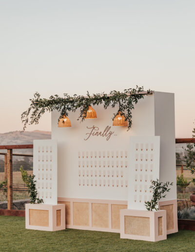 White display board decorated with greenery and hanging lights, featuring the word "Finally..." at the top. It stands on a grassy area with mountains visible in the background.