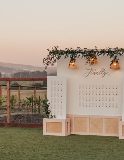 Decorative seating chart display at sunset, adorned with greenery and lights, with the word "Finally" at the top. A scenic landscape is visible in the background.