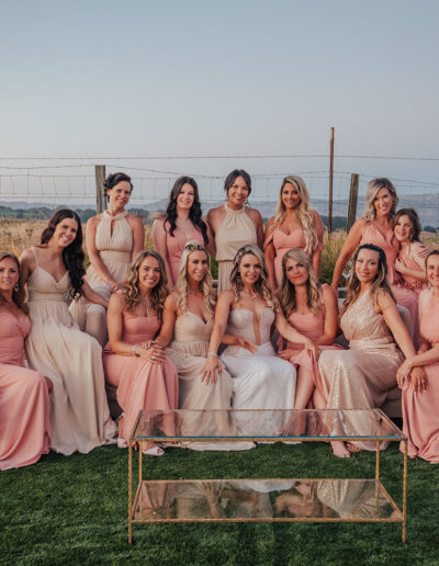 A group of women and a girl in pink and beige dresses sit and stand together outdoors. A stone wall and fence are in the background.