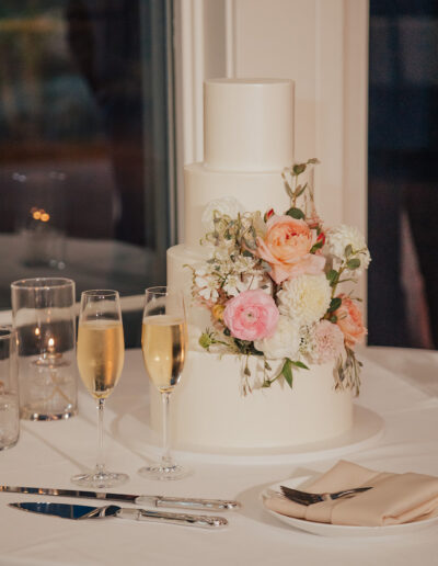 A three-tier white cake adorned with pink and white flowers next to two champagne flutes, candles, cutlery, and a folded napkin on a table.