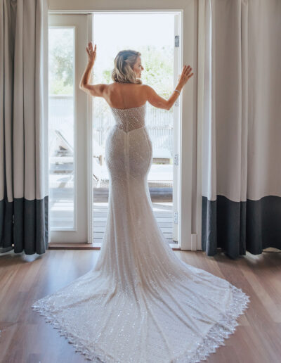 A woman in a sequin-embellished, strapless gown stands facing a glass door with curtains, her back to the camera.