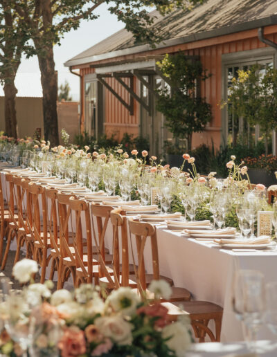 A long outdoor dining table is set with white tablecloths, wooden chairs, glassware, and floral centerpieces, next to a rustic building surrounded by greenery.