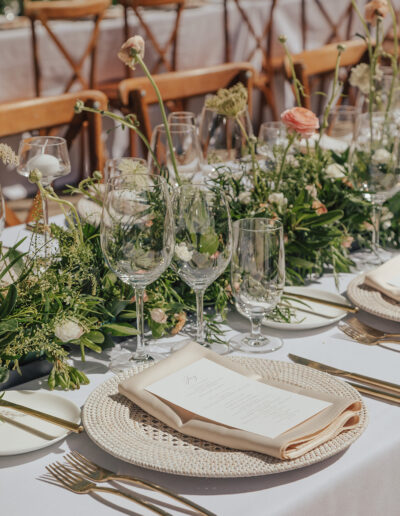 A table set for a formal outdoor event with white tablecloth, greenery centerpieces, wine glasses, plates, and silverware.