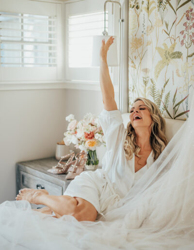 A woman in a white robe sits joyfully on a bed, reaching for a lamp. A bouquet and shoes are on the bedside table.