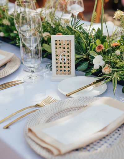 Elegant table setting with gold utensils, woven placemats, and white plates. A floral centerpiece runs along the middle, with a table number card displayed prominently.